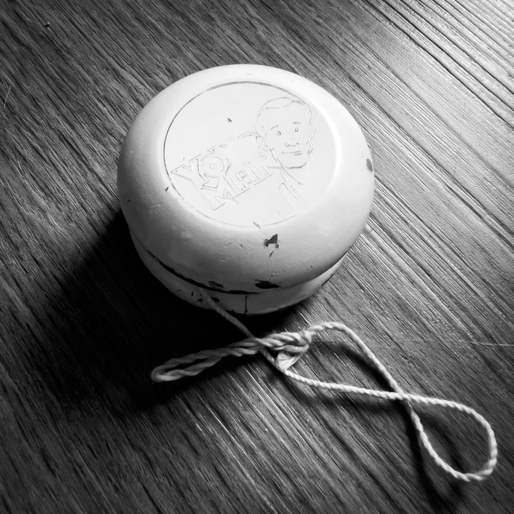 Black and white photo of a white yo-yo on a dark floor. Faded YoYo Man engraving face up. 