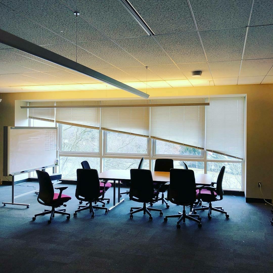 Distance photo of an empty conference table in front of windows. The blinds on the windows are at varying heights, their bottom edge lined up creating an arc across the windows.