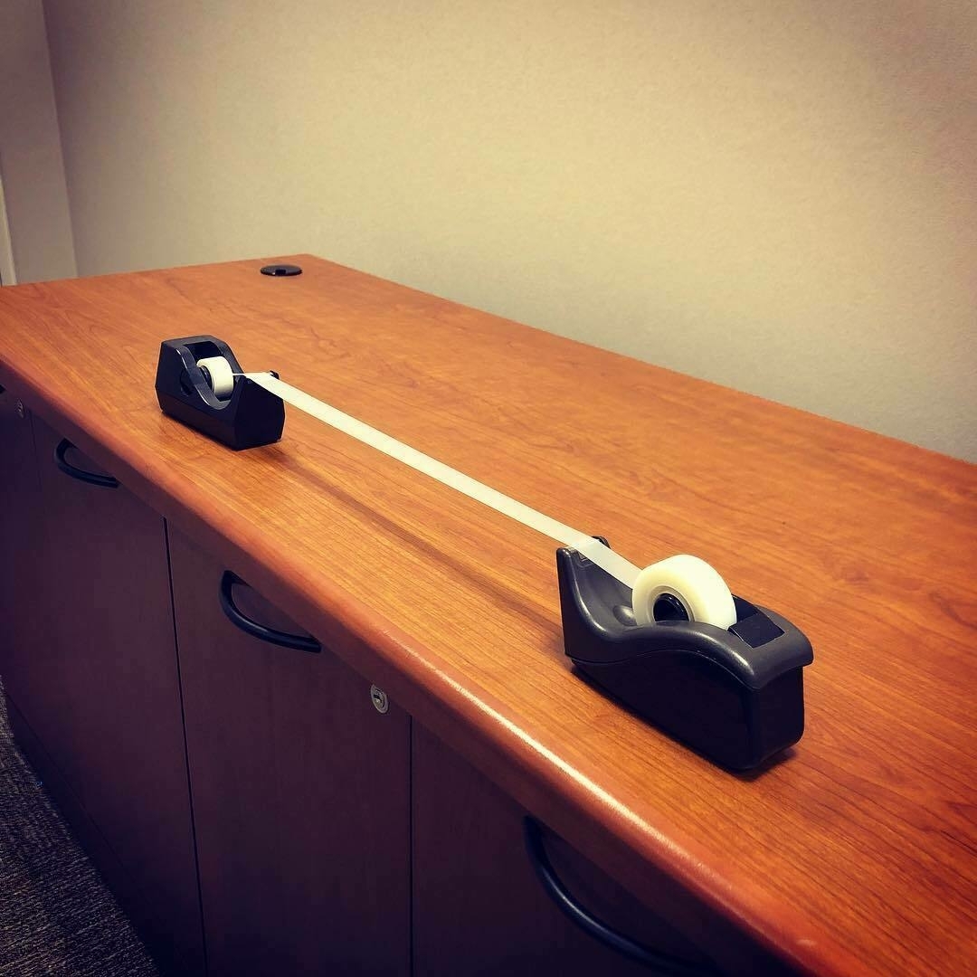 Two tape dispensers on a wooden desk, facing eachother with their tape connected and stretched several inches apart.